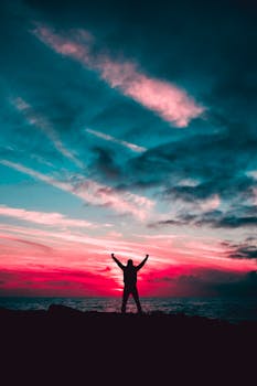 Person silhouetted against a vibrant sunset sky over the ocean, arms raised triumphantly.
